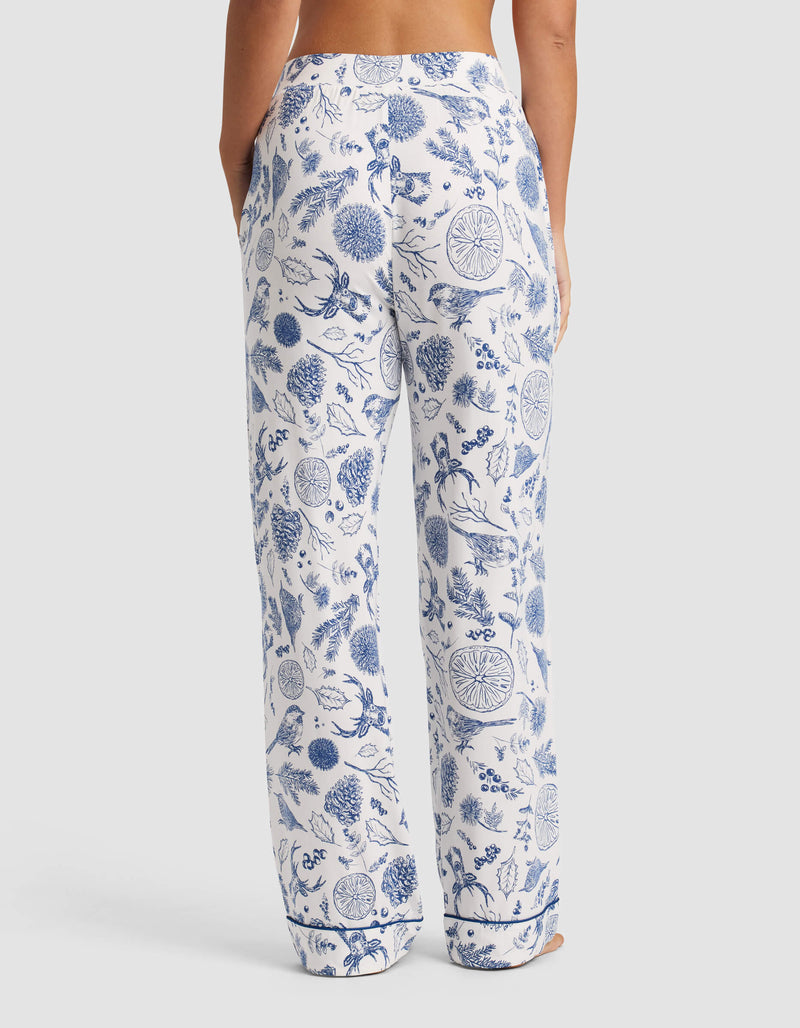 A person stands facing away, wearing Cozy Earth Women's Bamboo Stretch-Knit Pajama Pant in white with blue floral patterns. The relaxed-fit pants feature side pockets; the upper body is not fully visible.