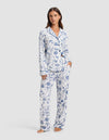 A woman stands barefoot wearing Cozy Earth's Women's Bamboo Stretch-Knit Pajama Pant, featuring a white background with blue botanical print and paired with a matching long-sleeve button-up top.|Color:Winter Toile Pacific