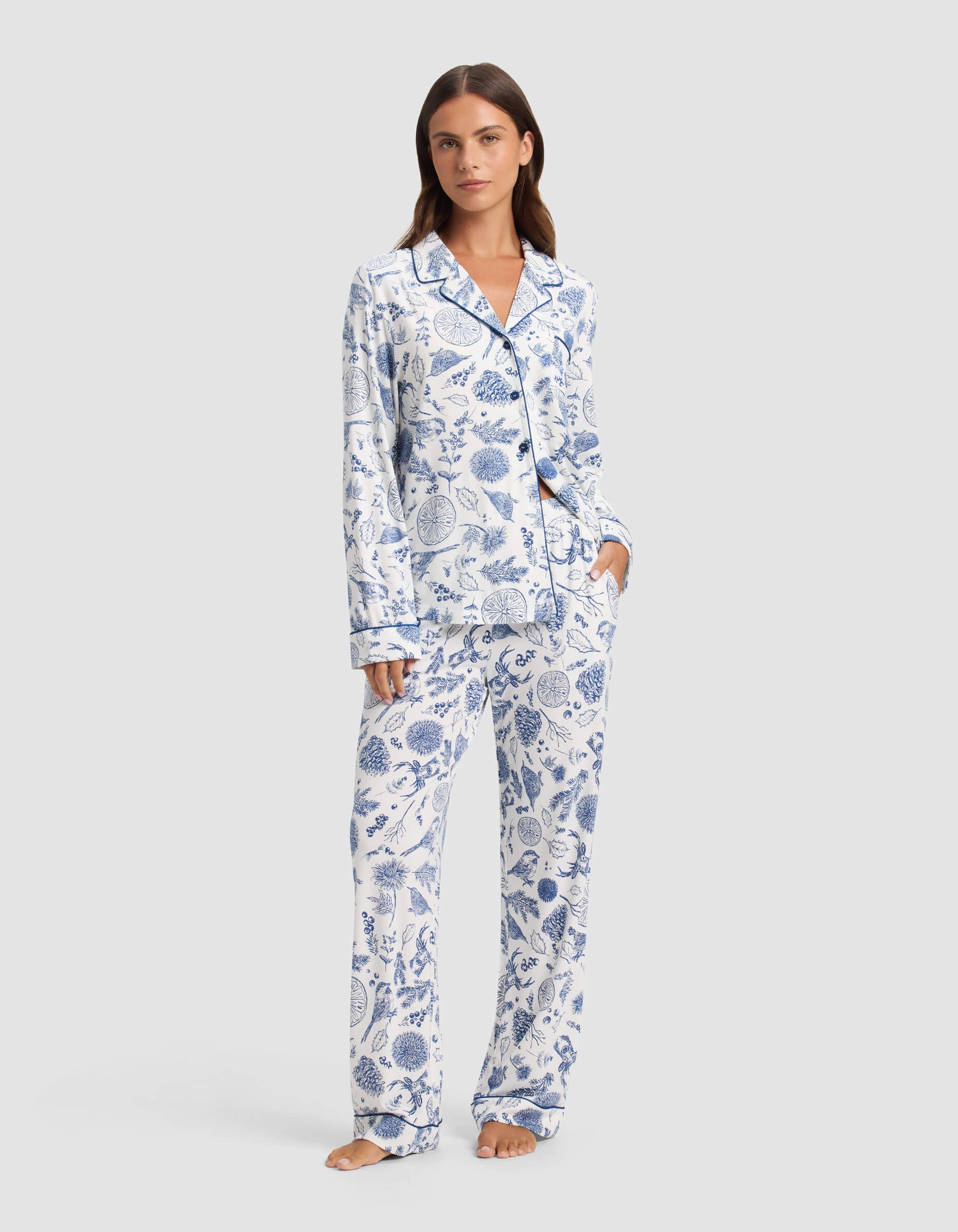 A woman stands barefoot wearing Cozy Earth's Women's Bamboo Stretch-Knit Pajama Pant, featuring a white background with blue botanical print and paired with a matching long-sleeve button-up top.