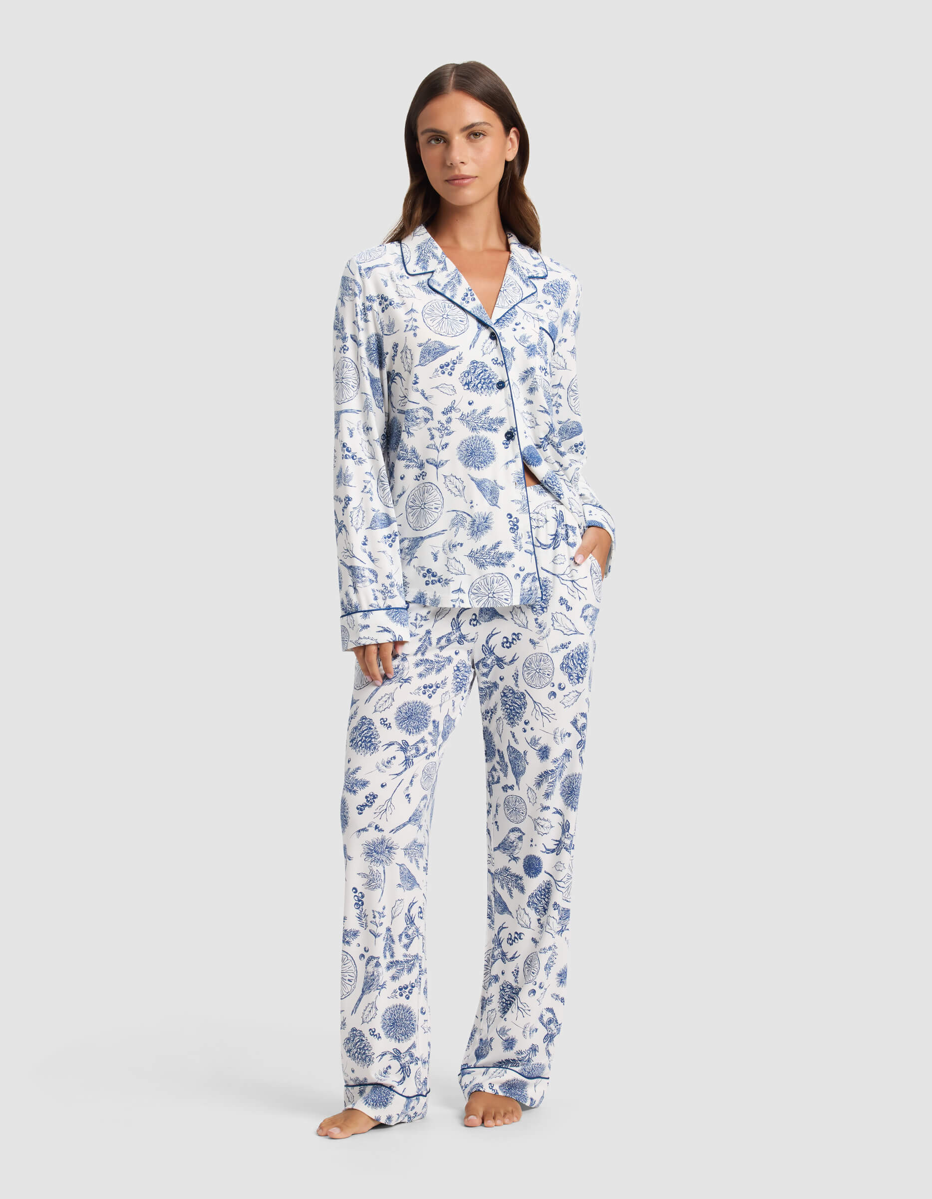 A woman stands barefoot wearing Cozy Earth's Women's Bamboo Stretch-Knit Pajama Pant, featuring a white background with blue botanical print and paired with a matching long-sleeve button-up top.|Color:Winter Toile Pacific