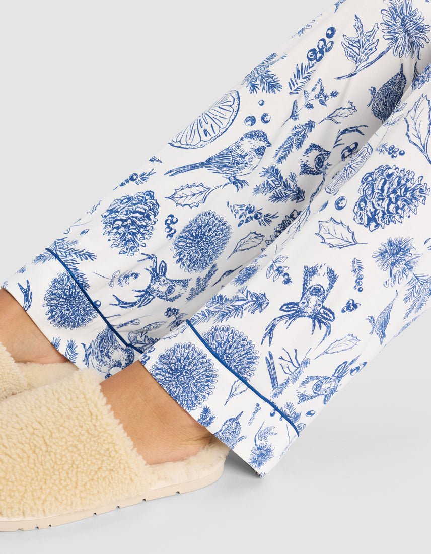 Close-up of a person wearing cream-colored slippers and Cozy Earth Women's Bamboo Stretch-Knit Pajama Pant, featuring a blue nature print with birds, leaves, pinecones, and acorns, set against a light background.