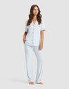 A woman with long brown hair and a relaxed expression stands barefoot, wearing the Cozy Earth Women's Bamboo Stretch-Knit Long Sleeve Pajama Set. The outfit features a button-up top and long pants in a soft, stretchy fabric. |Color:Market Stripe in Lake