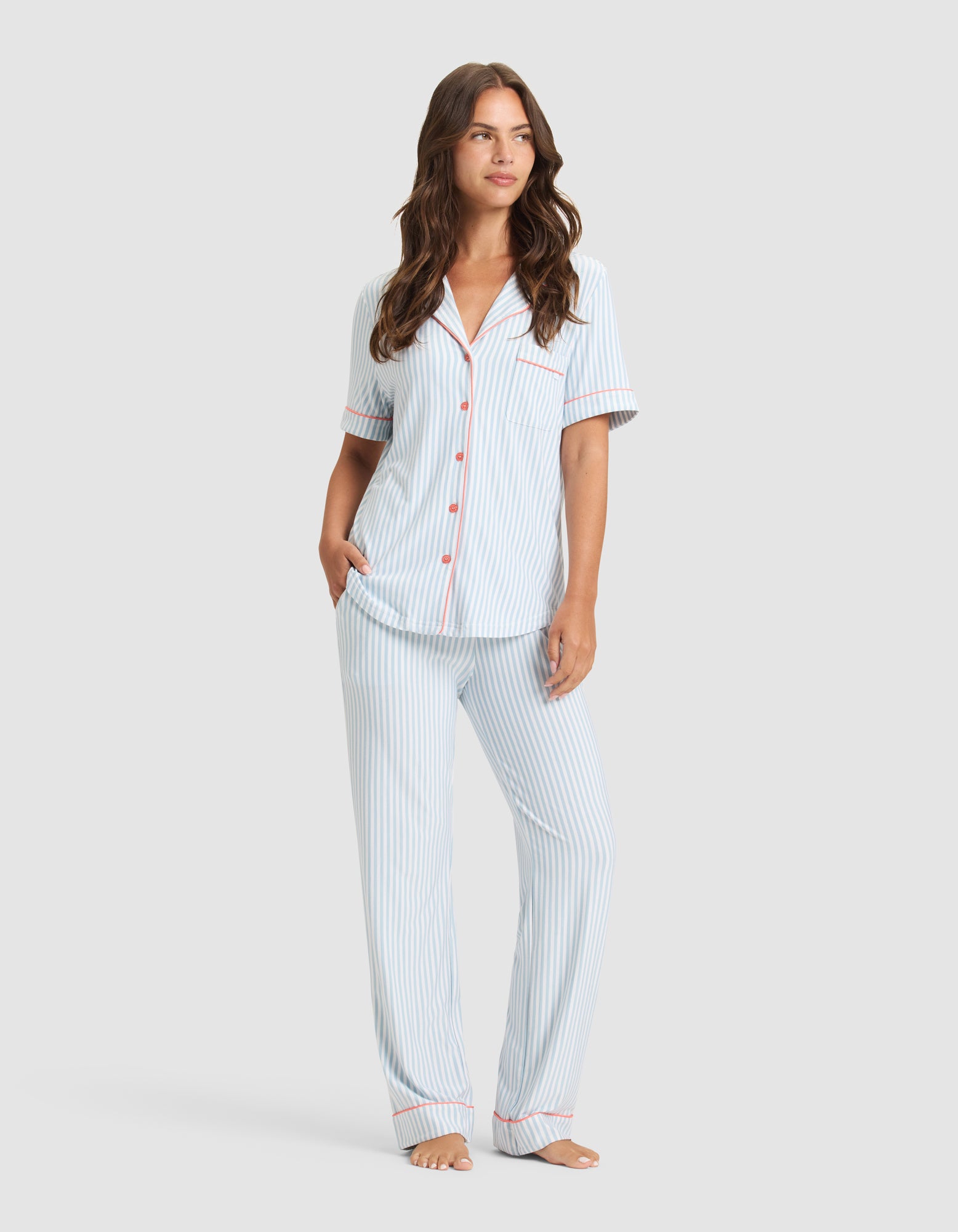 A woman with long brown hair and a relaxed expression stands barefoot, wearing the Cozy Earth Women's Bamboo Stretch-Knit Long Sleeve Pajama Set. The outfit features a button-up top and long pants in a soft, stretchy fabric. |Color:Market Stripe in Lake