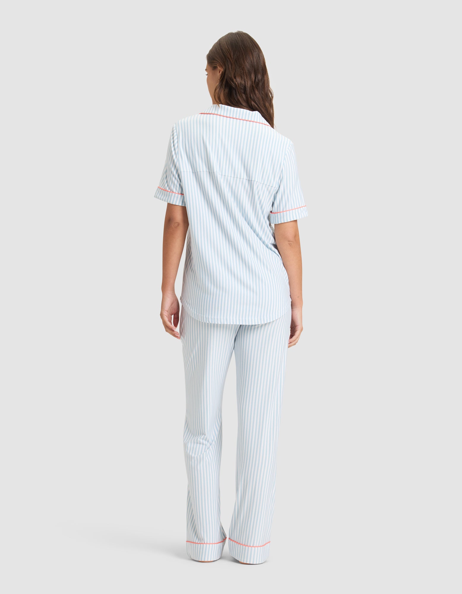 A woman stands facing away in the Cozy Earth Women's Bamboo Stretch-Knit Long Sleeve Pajama Set, featuring light blue and white vertical stripes with pink piping. The background is a plain light gray.
