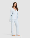 A woman models the Cozy Earth Women's Bamboo Stretch-Knit Long Sleeve Pajama Set—light blue and white stripes with red piping, button-up top, and matching pants—against a plain light background. |Color:Market Stripe in Lake