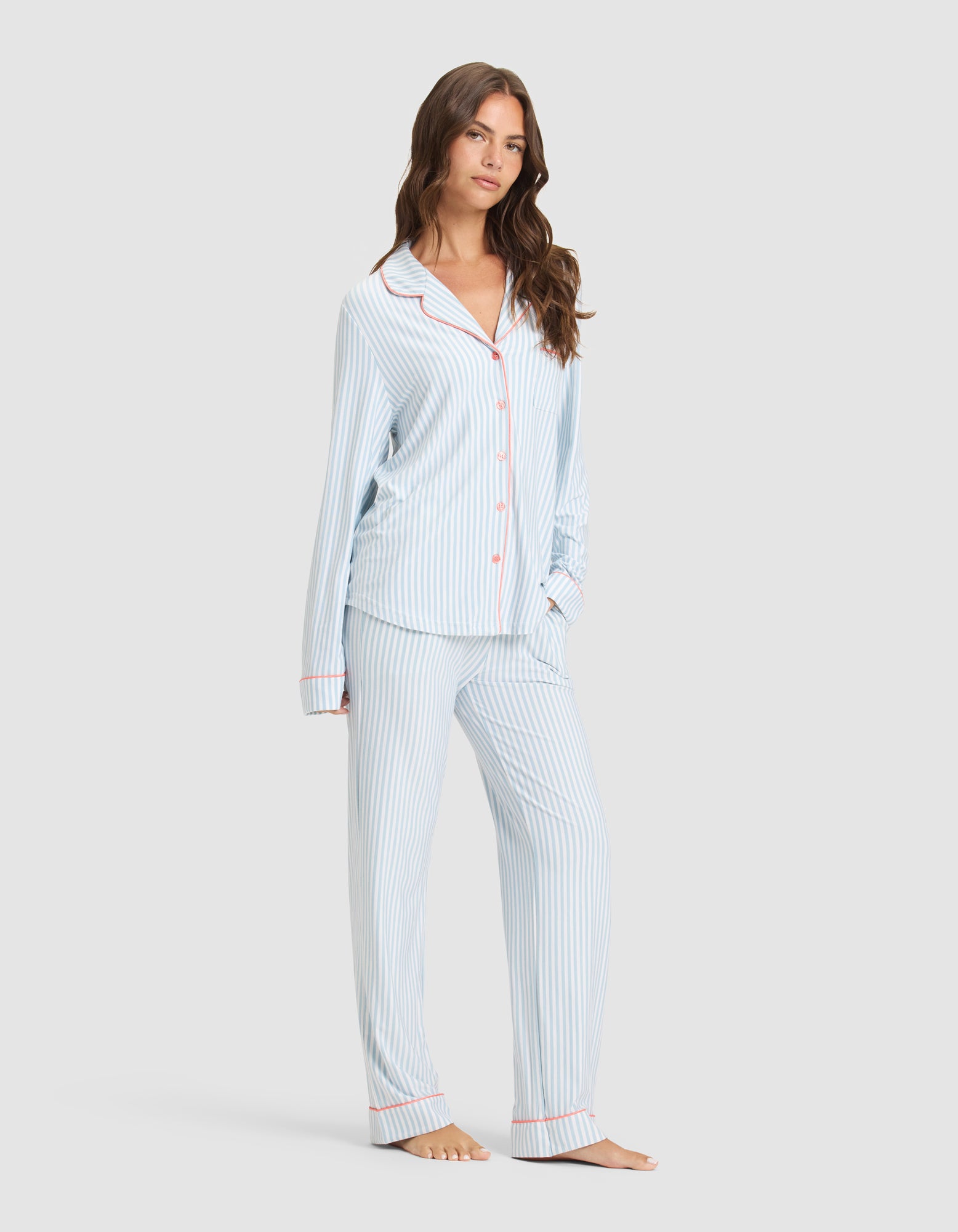 A woman models the Cozy Earth Women's Bamboo Stretch-Knit Long Sleeve Pajama Set—light blue and white stripes with red piping, button-up top, and matching pants—against a plain light background. |Color:Market Stripe in Lake