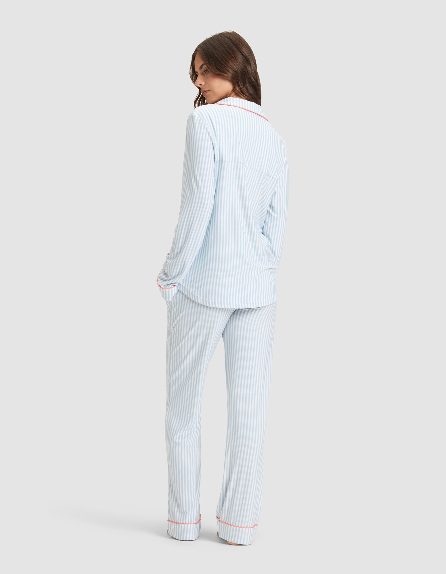 A woman smiles over her shoulder, hands in pockets, wearing Cozy Earth's Women's Bamboo Stretch-Knit Long Sleeve Pajama Set in light blue and white stripes with red piping. She stands against a plain light gray background. 
