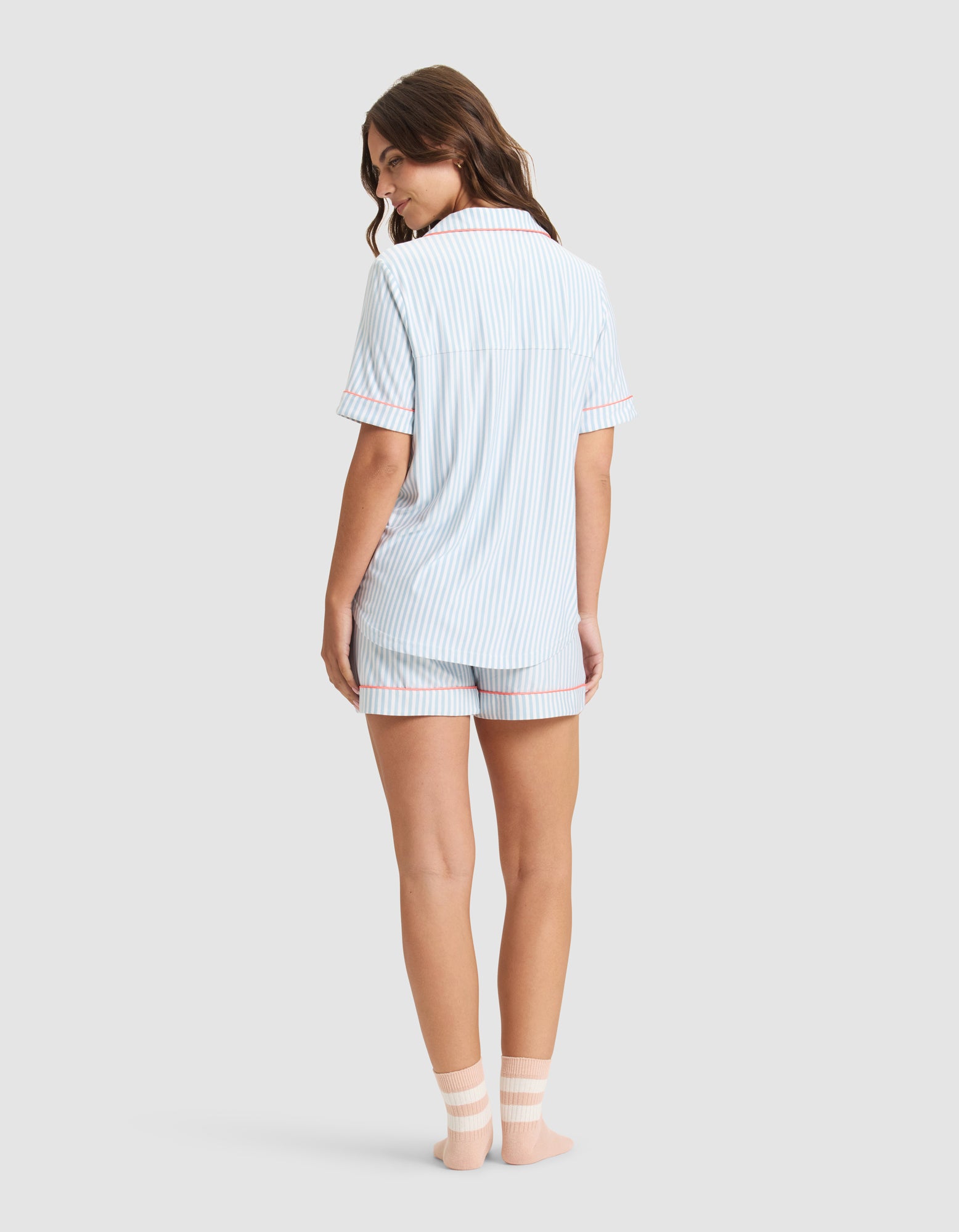 A woman faces away in Cozy Earth's Women's Bamboo Stretch-Knit Short Sleeve Pajama Set—light blue and white stripes—with matching shorts and top, plus white ankle socks, against a plain light gray background. 