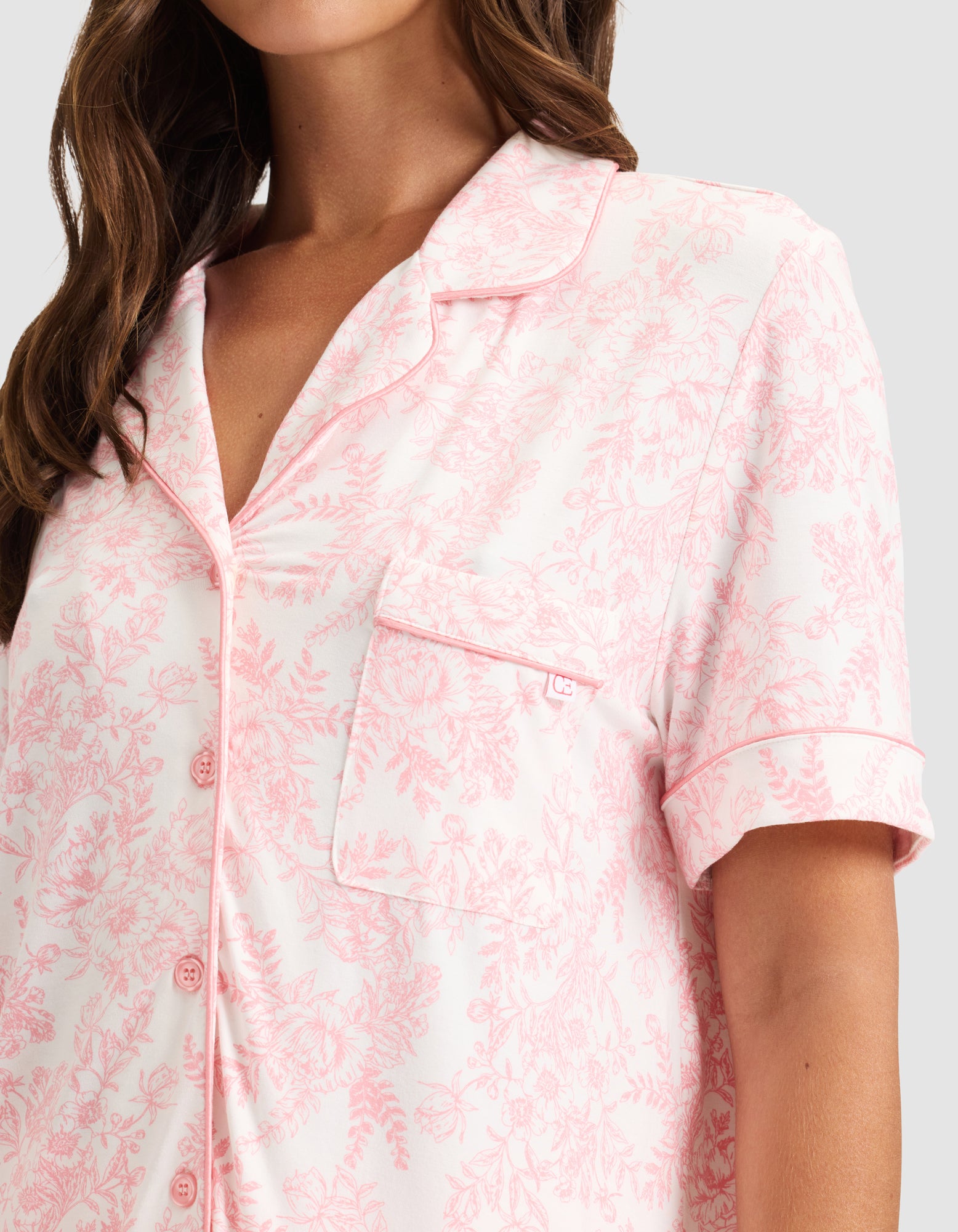 A woman wears HIDE's Women's Bamboo Stretch-Knit Short Sleeve Pajama Top in light pink with a floral pattern, notched collar, chest pocket, front buttons, and pink piping. Only her upper body is visible. 