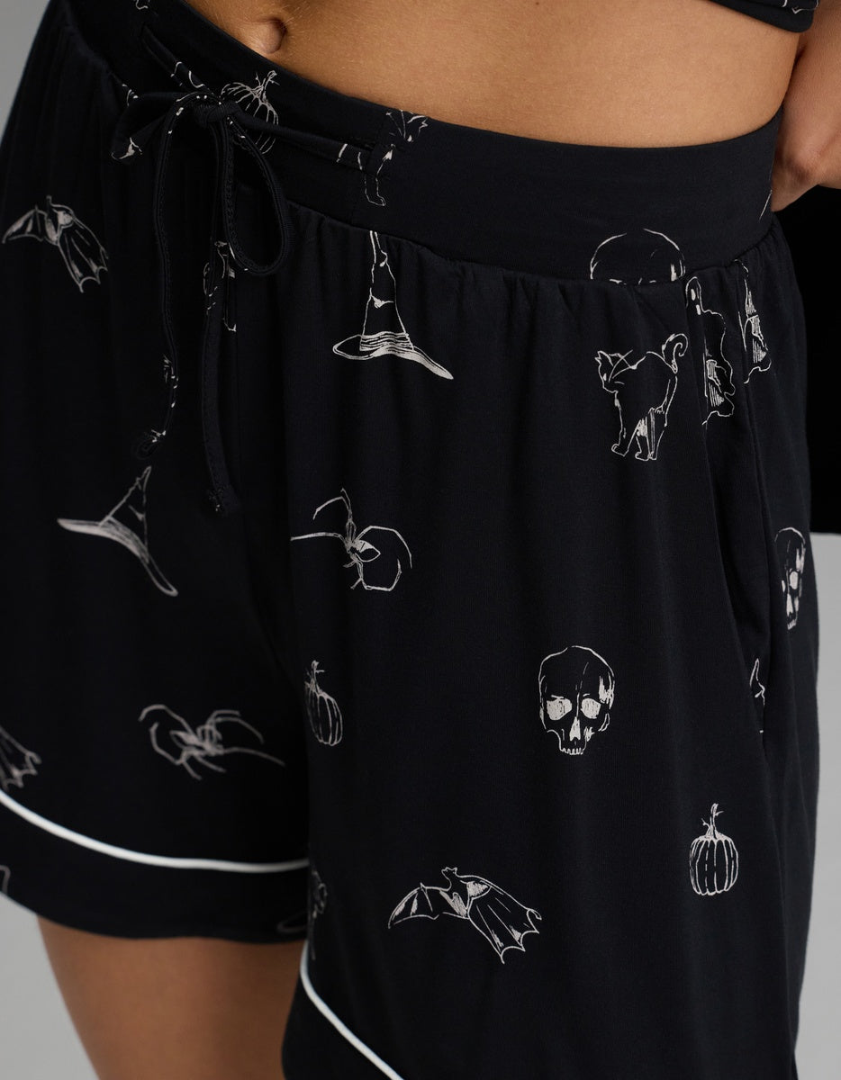 Close-up of the Women’s Bamboo Stretch-Knit Short Sleeve Pajama Set by Cozy Earth, featuring black shorts with white Halloween-themed prints—skull, bat, pumpkin, witch hat, and cat—accented by white piping details.