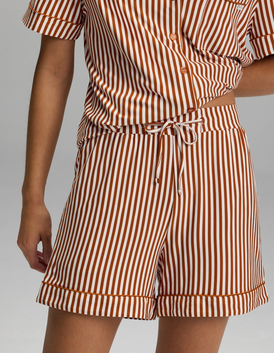 A person is shown from shoulder to mid-thigh wearing the Cozy Earth Women’s Bamboo Stretch-Knit Pajama Short, part of a matching short-sleeve button-up shirt and shorts set featuring vertical white and brown stripes.