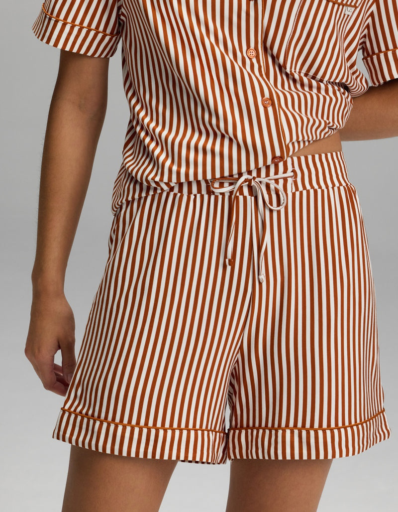 A person is shown from shoulder to mid-thigh wearing the Cozy Earth Women’s Bamboo Stretch-Knit Pajama Short, part of a matching short-sleeve button-up shirt and shorts set featuring vertical white and brown stripes.