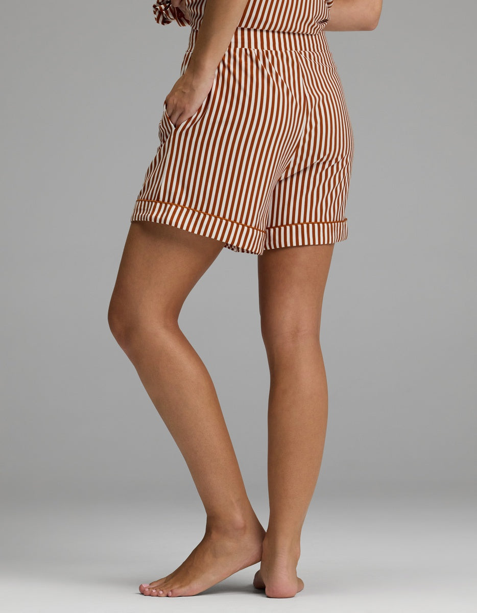 A person standing barefoot on one leg wears Cozy Earth Women’s Bamboo Stretch-Knit Pajama Shorts in a brown and white vertical stripe pattern, with their upper body mostly out of frame against a plain gray background.