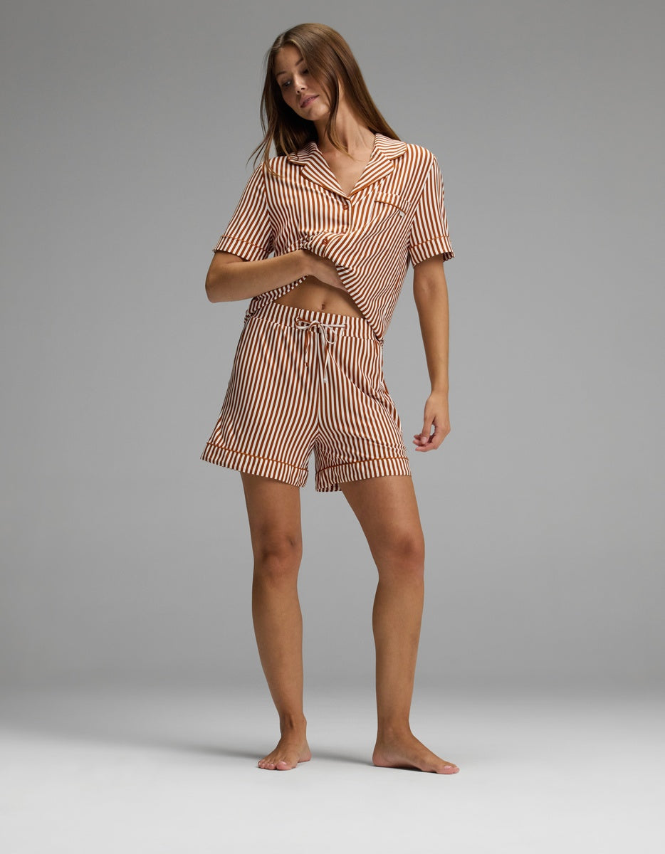 A woman stands barefoot on a gray background, wearing the Cozy Earth Women’s Bamboo Stretch-Knit Pajama Short set, which features a matching short-sleeve, button-up striped shirt and shorts. She is lifting one side of her shirt slightly and smiling, with her long hair down.