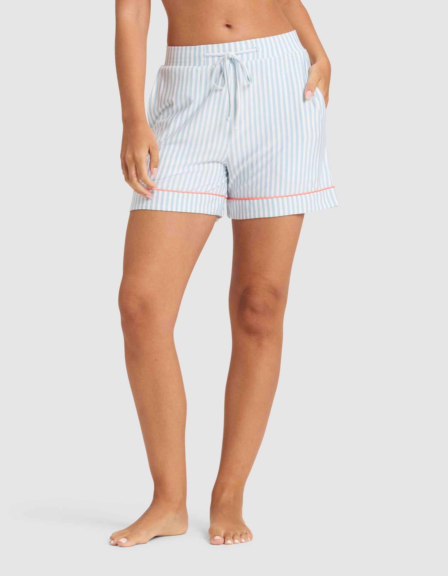 A person stands barefoot against a plain background wearing HIDE Women's Bamboo Stretch-Knit Pajama Shorts, which feature light blue and white stripes, a drawstring waist, and red trim at the hem. 