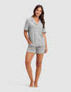 A woman smiles barefoot in Cozy Earth's Women's Bamboo Stretch-Knit Short Sleeve Pajama Set—featuring a button-up top and matching shorts in light grey—against a plain white background. |Color:Grey