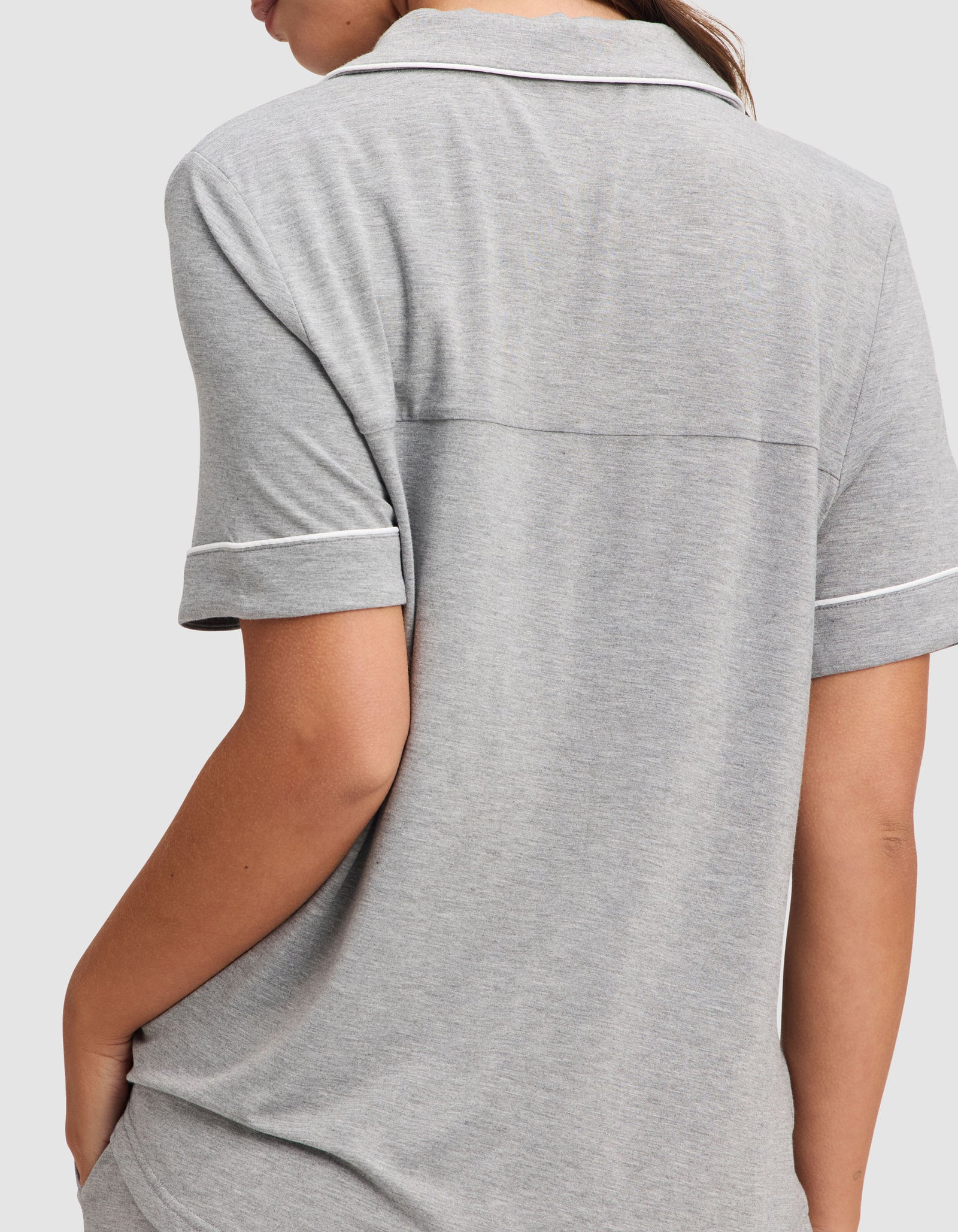 A woman is shown from the back wearing the Cozy Earth Women's Bamboo Stretch-Knit Short Sleeve Pajama Set in light gray, featuring a collared top with white trim and short sleeves, with her left arm bent and hand in her pocket. 