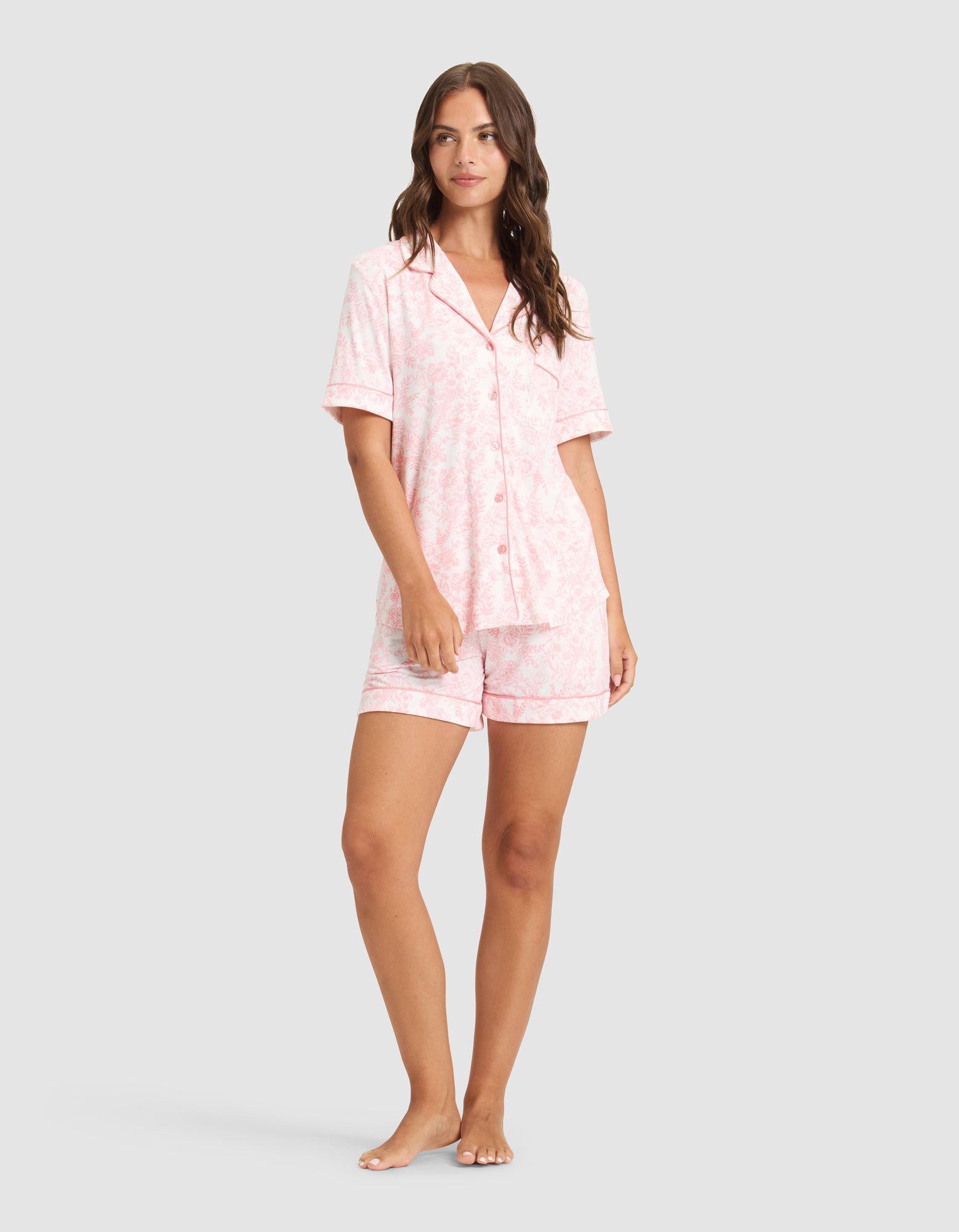 A woman stands barefoot wearing HIDE's Women's Bamboo Stretch-Knit Pajama Shorts and a matching light pink, short-sleeve button-up top with a subtle pattern, set against a plain light gray background. 