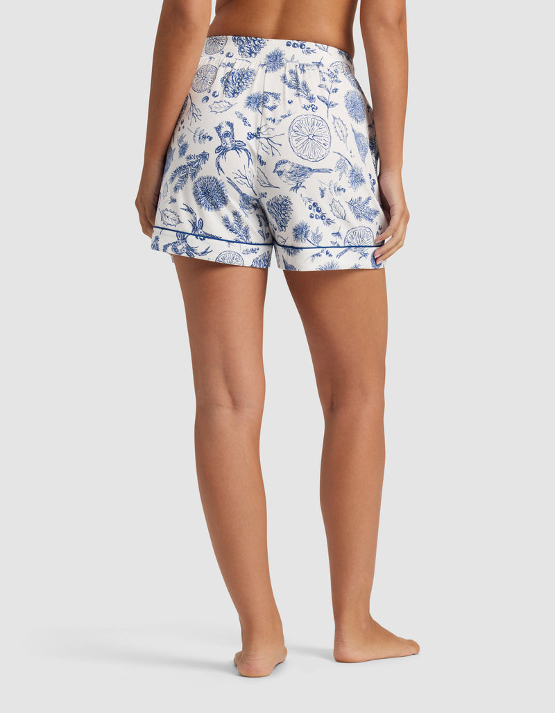 A woman stands barefoot, shown from the back, wearing Cozy Earth Women's Bamboo Stretch-Knit Pajama Shorts in a blue botanical and floral print. These shorts feature an elastic waistband and a relaxed fit.
