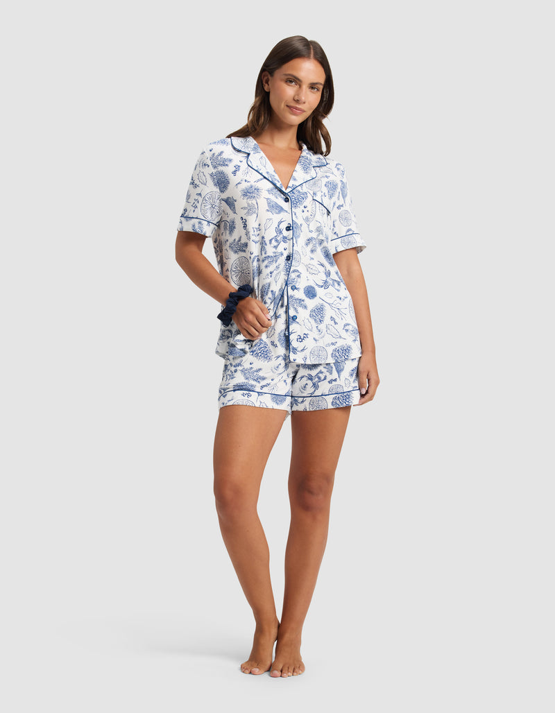 A woman smiles barefoot in the Cozy Earth Women's Bamboo Stretch-Knit Pajama Short set, featuring a blue and white floral short-sleeve button-up top with matching shorts. She holds a coordinating scrunchie against a plain light gray background.
