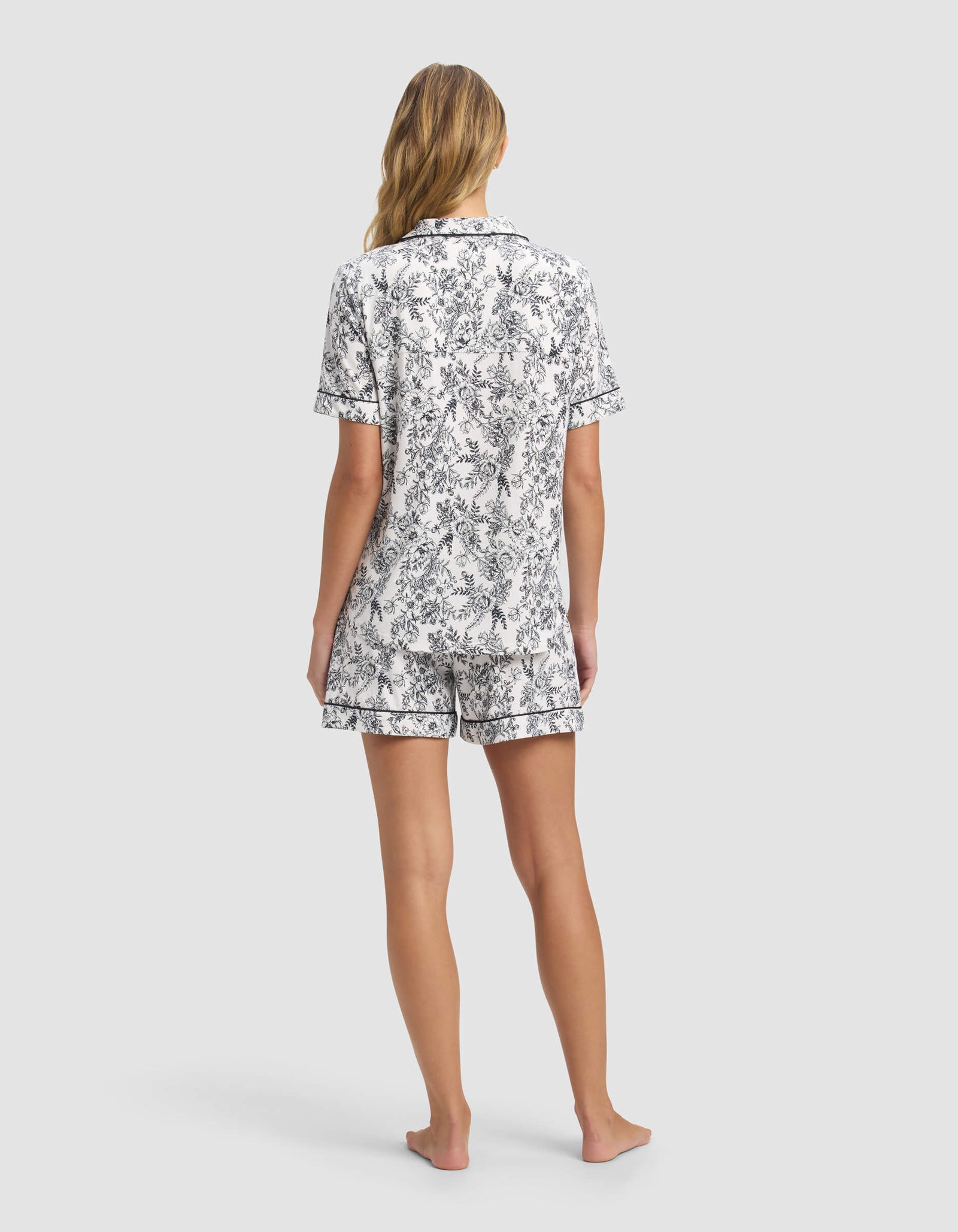 A woman with long, wavy blonde hair stands barefoot, facing away, wearing Cozy Earth’s Women's Bamboo Stretch-Knit Short Sleeve Pajama Set in a black and white floral print. The background is plain light gray.