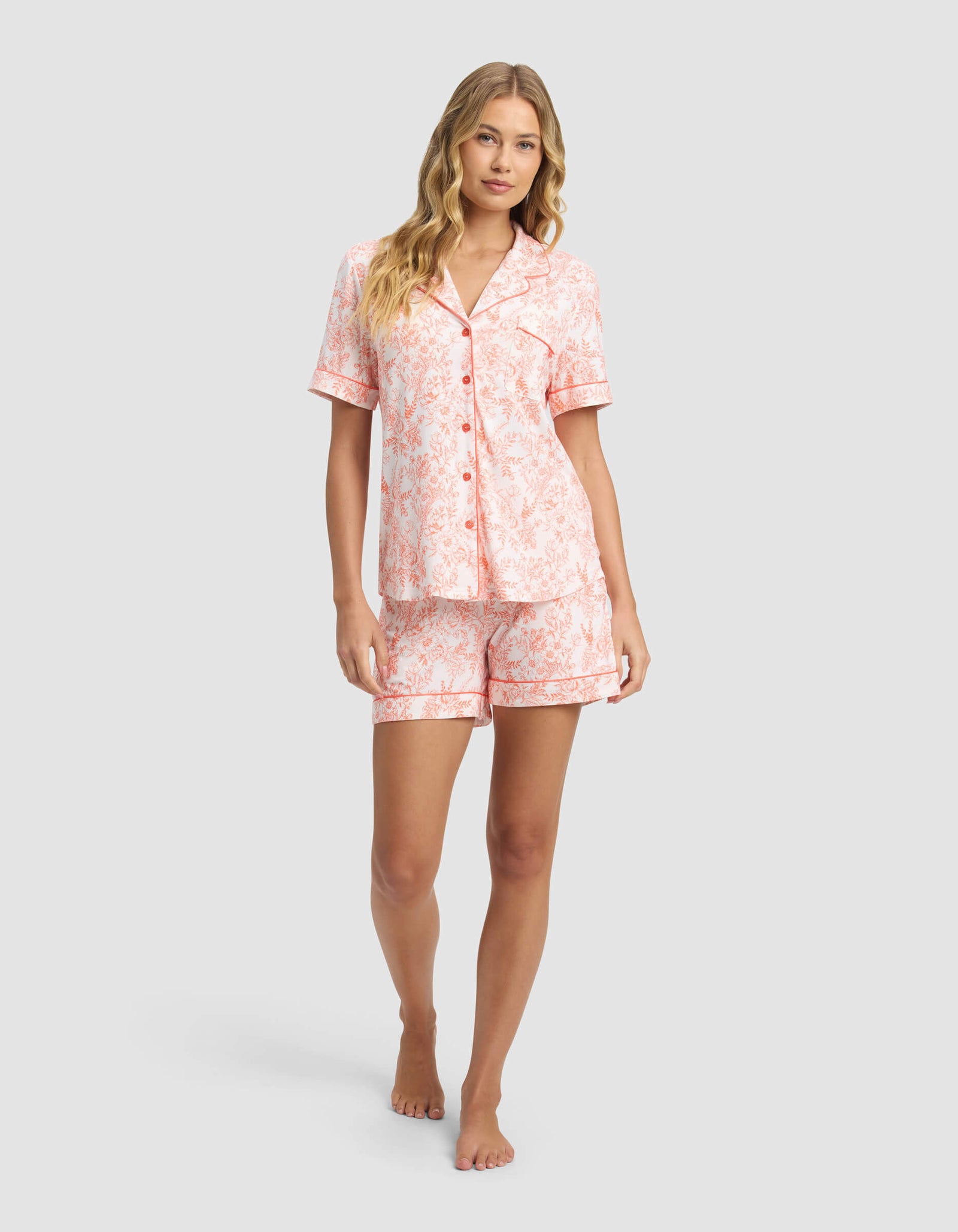 A woman stands barefoot against a light gray background wearing the Cozy Earth Women's Bamboo Stretch-Knit Short Sleeve Pajama Set, featuring a matching button-up top and shorts with a light pink and white floral pattern.