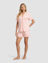 A woman stands barefoot against a light gray background, wearing Cozy Earth’s Women's Bamboo Stretch-Knit Short Sleeve Pajama Set in pink and white with a collared button-up top, front pocket, and floral shorts.|Color:Melon Toile