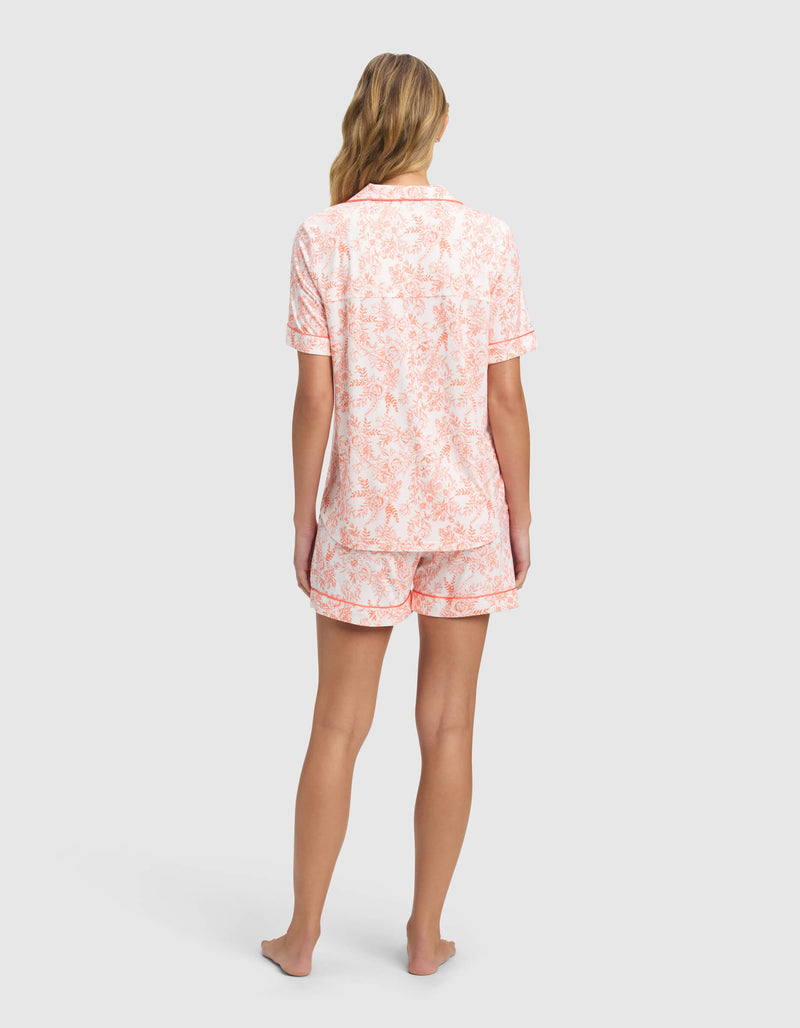 A woman with long, wavy blonde hair stands barefoot facing away, wearing the Cozy Earth Women's Bamboo Stretch-Knit Short Sleeve Pajama Set in white with a light pink floral pattern.