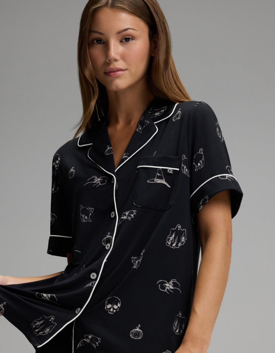 A woman wearing the Cozy Earth Women’s Bamboo Stretch-Knit Short Sleeve Pajama Set featuring black fabric with white Halloween-themed prints—pumpkins, ghosts, bats, and skulls—stands against a plain gray background.