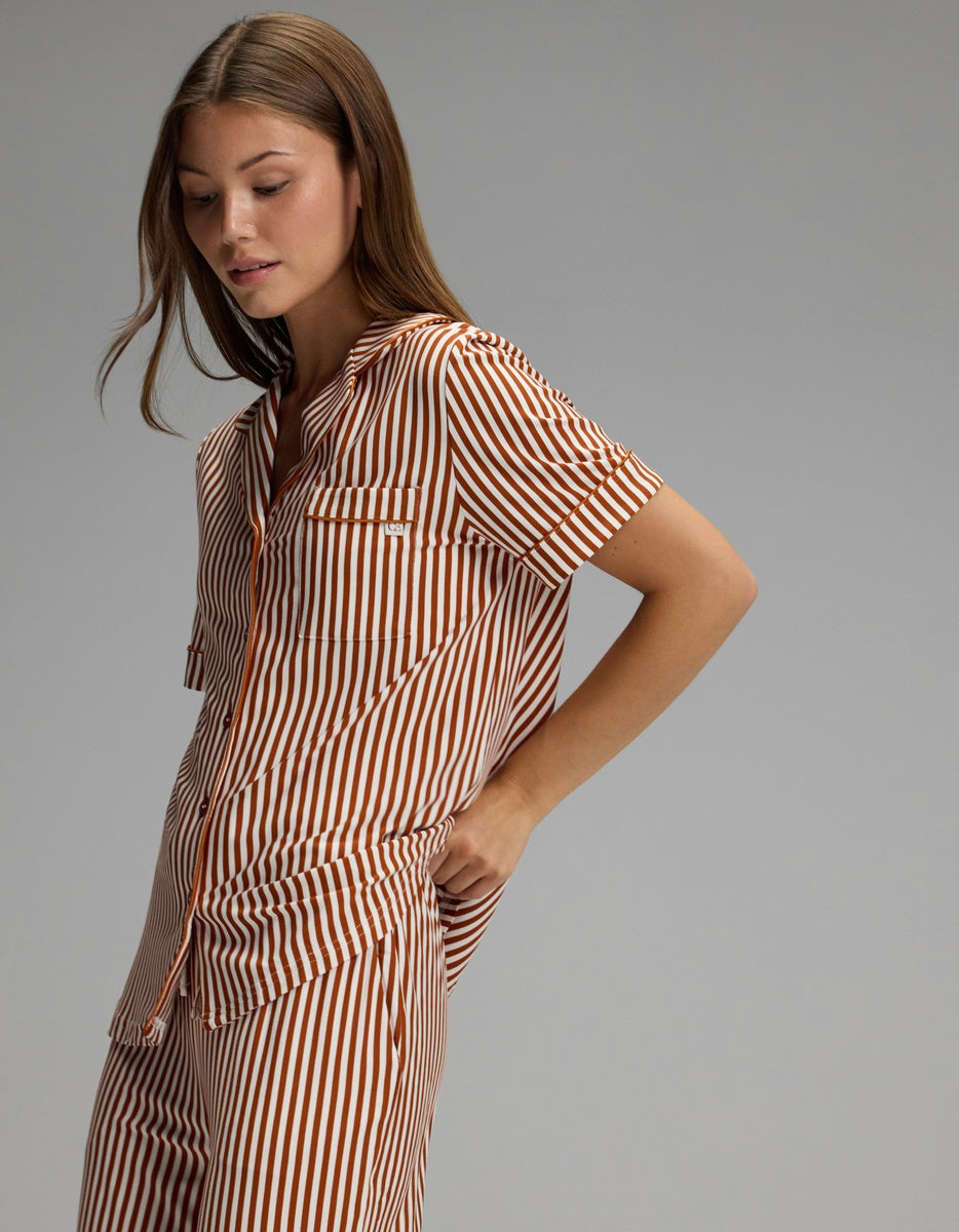 A person wears the Cozy Earth Women’s Bamboo Stretch-Knit Short Sleeve Pajama Top in brown and white stripes, standing against a plain gray background with one hand in their pocket and looking down.