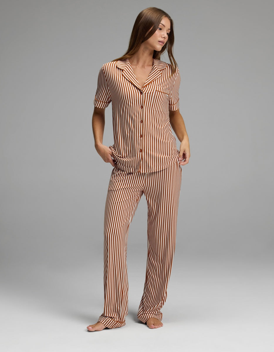 A person stands barefoot against a plain gray background, wearing Cozy Earth’s Women’s Bamboo Stretch-Knit Short Sleeve Pajama Top, which features vertical orange and white stripes, short sleeves, buttons down the front, and is paired with matching long pants. The person’s hands are in their pockets.