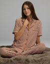 A woman with long brown hair sits cross-legged on a plush gray blanket, calmly looking toward the camera while wearing the Cozy Earth Women’s Bamboo Stretch-Knit Short Sleeve Pajama Top in brown and white stripes.|Color:Market Stripe in Cinnamon