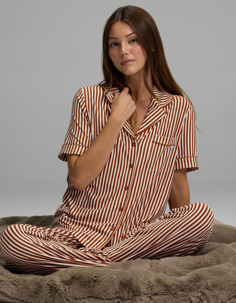 A woman with long brown hair sits cross-legged on a plush gray blanket, calmly looking toward the camera while wearing the Cozy Earth Women’s Bamboo Stretch-Knit Short Sleeve Pajama Top in brown and white stripes.