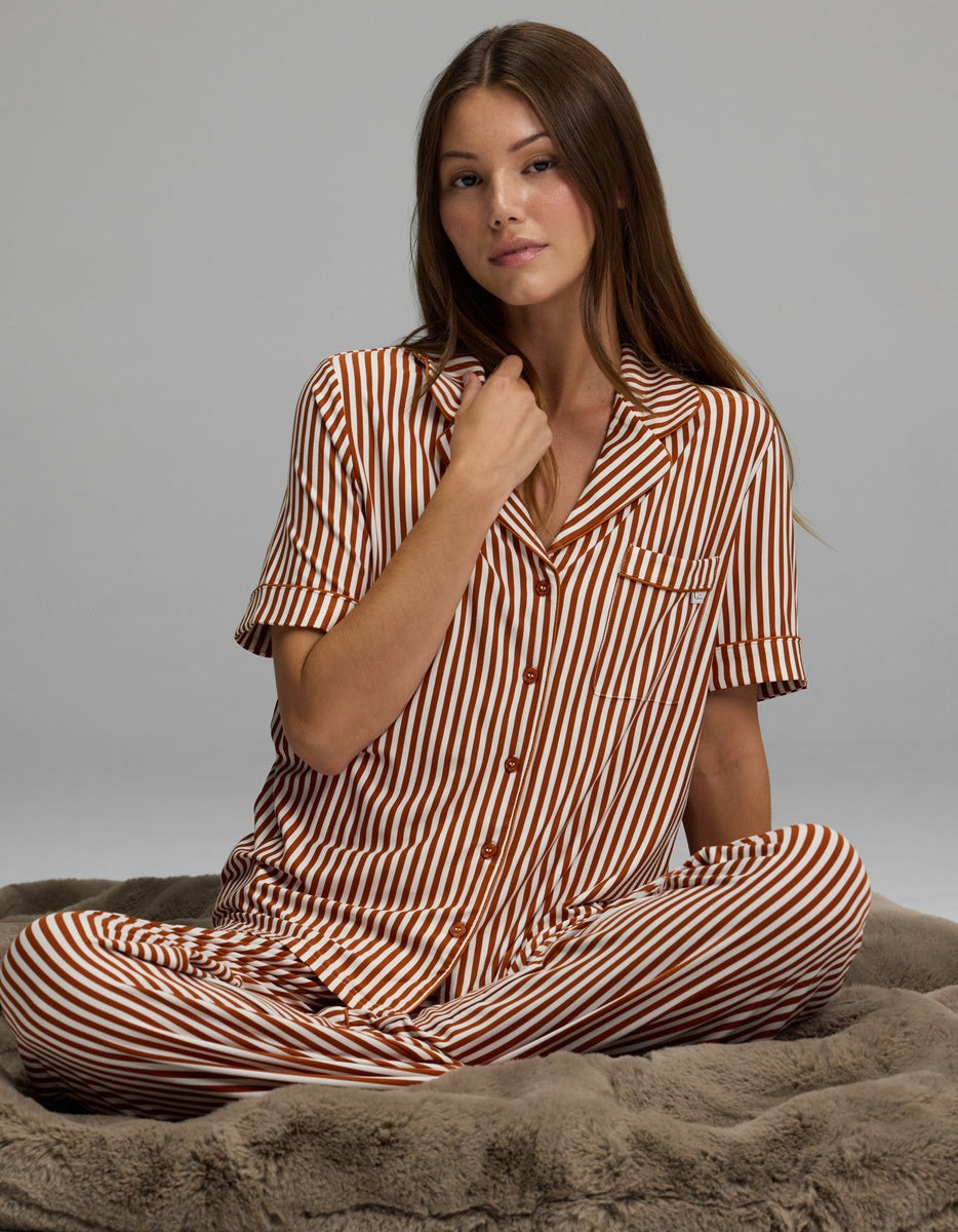 A woman with long brown hair sits cross-legged on a plush gray blanket, calmly looking toward the camera while wearing the Cozy Earth Women’s Bamboo Stretch-Knit Short Sleeve Pajama Top in brown and white stripes.|Color:Market Stripe in Cinnamon