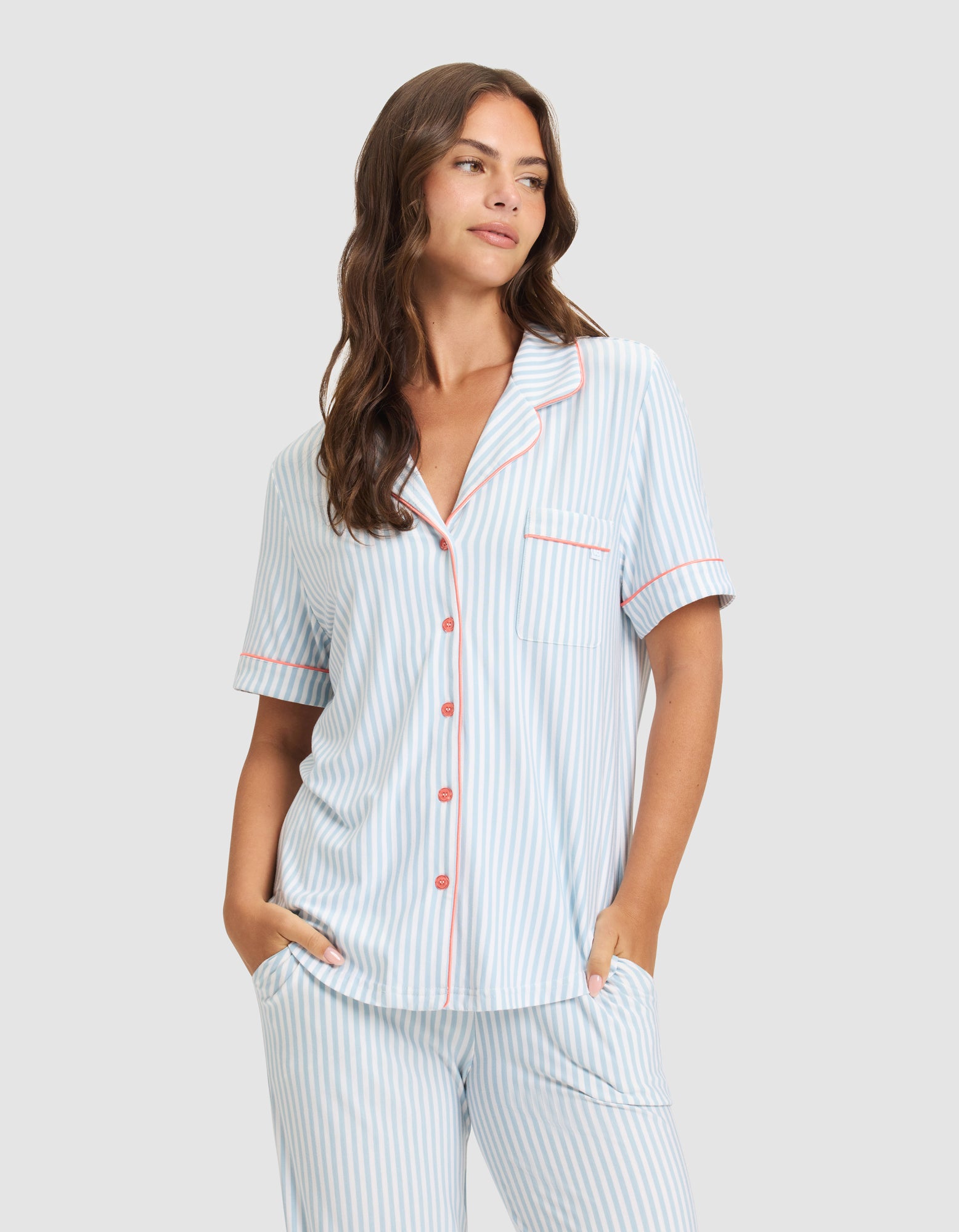 A woman wearing the HIDE Women's Bamboo Stretch-Knit Short Sleeve Pajama Top in light blue and white stripes with pink trim and front buttons stands against a plain light gray background, hands in her pockets. 