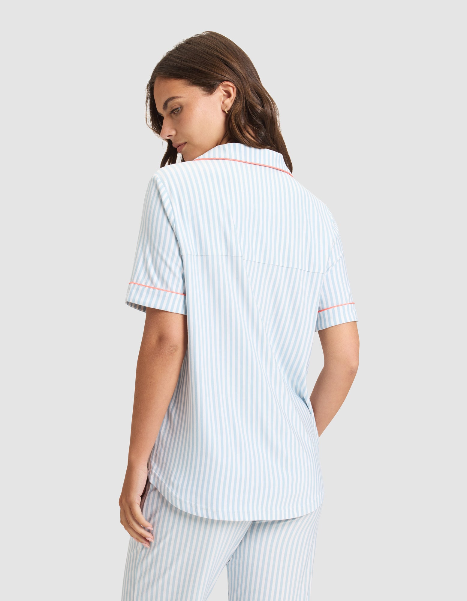 A woman with long brown hair wears the HIDE Women's Bamboo Stretch-Knit Short Sleeve Pajama Top in light blue and white stripes with pink trim. She stands with her back to the camera, looking over her shoulder against a plain light gray background. 