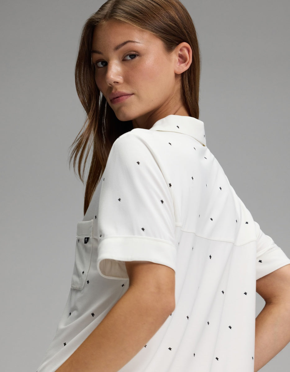 A person with long brown hair looks over their shoulder against a plain gray background, wearing the Cozy Earth Women’s Bamboo Stretch-Knit Short Sleeve Pajama Set in white with small black dots.