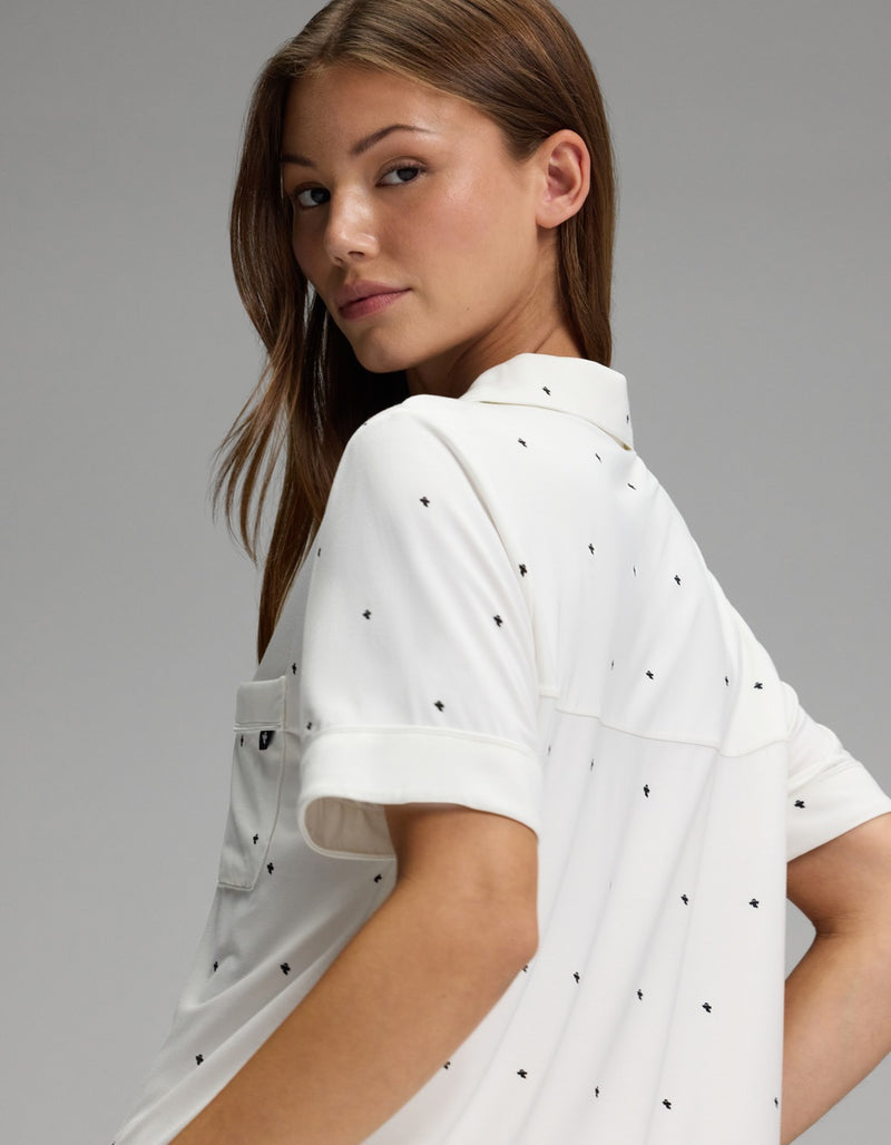 A person with long brown hair looks over their shoulder against a plain gray background, wearing the Cozy Earth Women’s Bamboo Stretch-Knit Short Sleeve Pajama Set in white with small black dots.