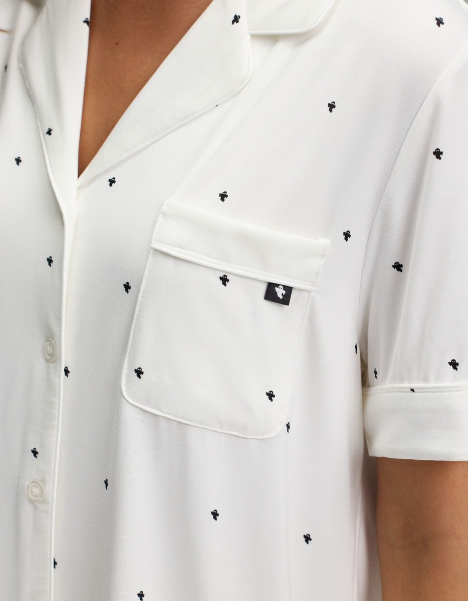 Close-up of the Cozy Earth Women’s Bamboo Stretch-Knit Short Sleeve Pajama Set in white, featuring a notched collar, chest pocket with black tag, and subtle small black embroidered designs. Part of the wearer’s neck and arm is visible.