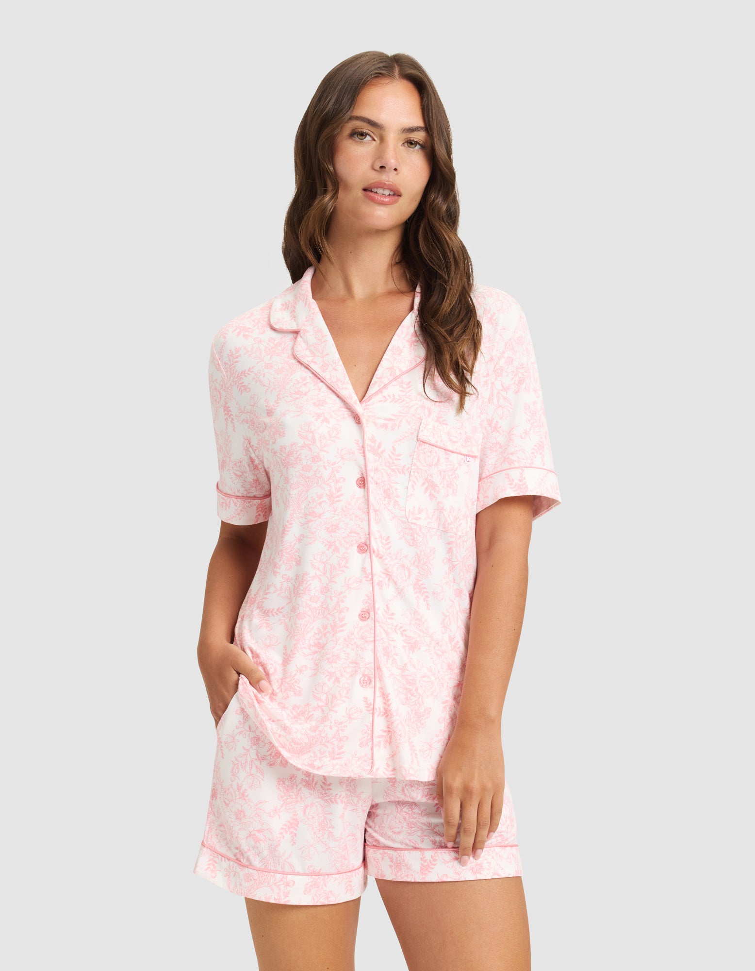 A woman with long brown hair models the HIDE Women's Bamboo Stretch-Knit Short Sleeve Pajama Top in light pink, featuring a subtle floral pattern, button-up front, and matching shorts, standing against a plain light background. 