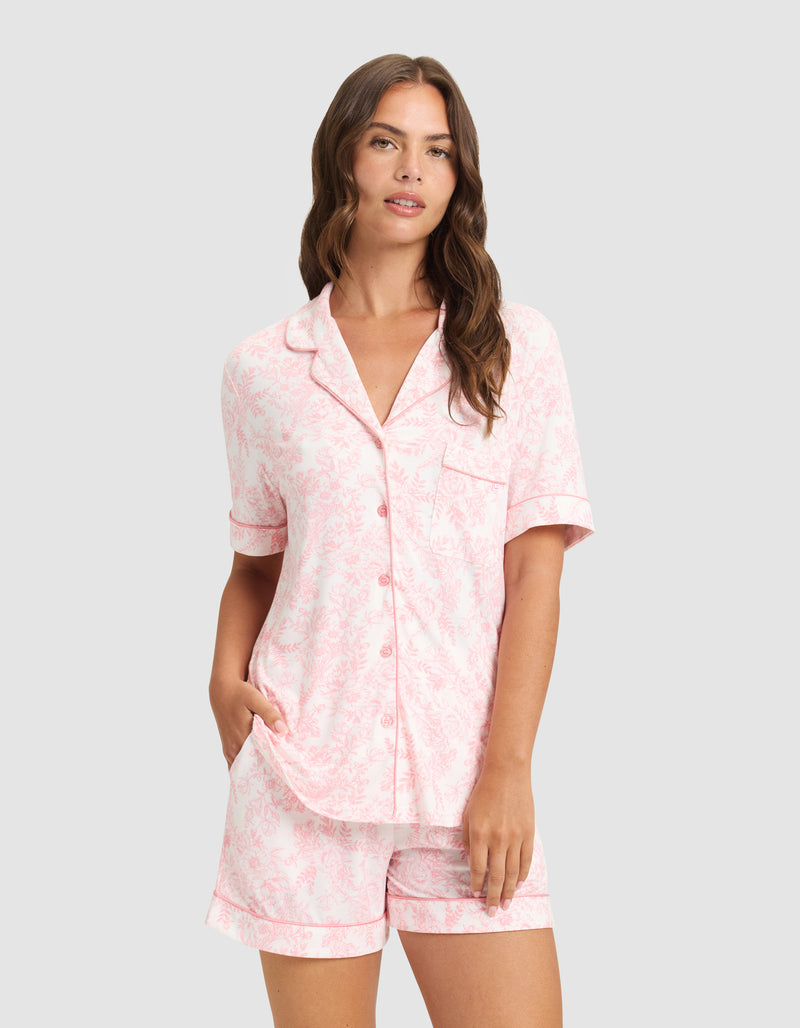 A woman with long brown hair models the HIDE Women's Bamboo Stretch-Knit Short Sleeve Pajama Top in light pink, featuring a subtle floral pattern, button-up front, and matching shorts, standing against a plain light background.