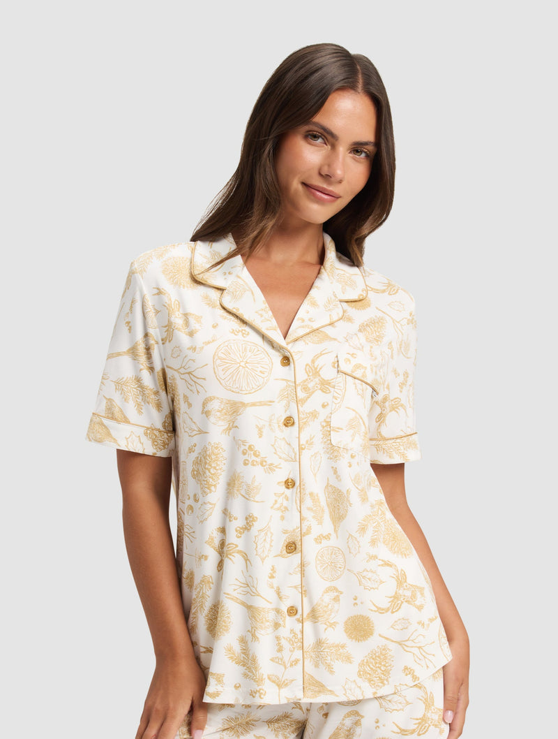 A woman with long brown hair models the Cozy Earth Women's Bamboo Stretch-Knit Short Sleeve Pajama Top, part of a white pajama set with gold botanical prints. She pairs it with matching shorts and smiles softly against a plain light background.