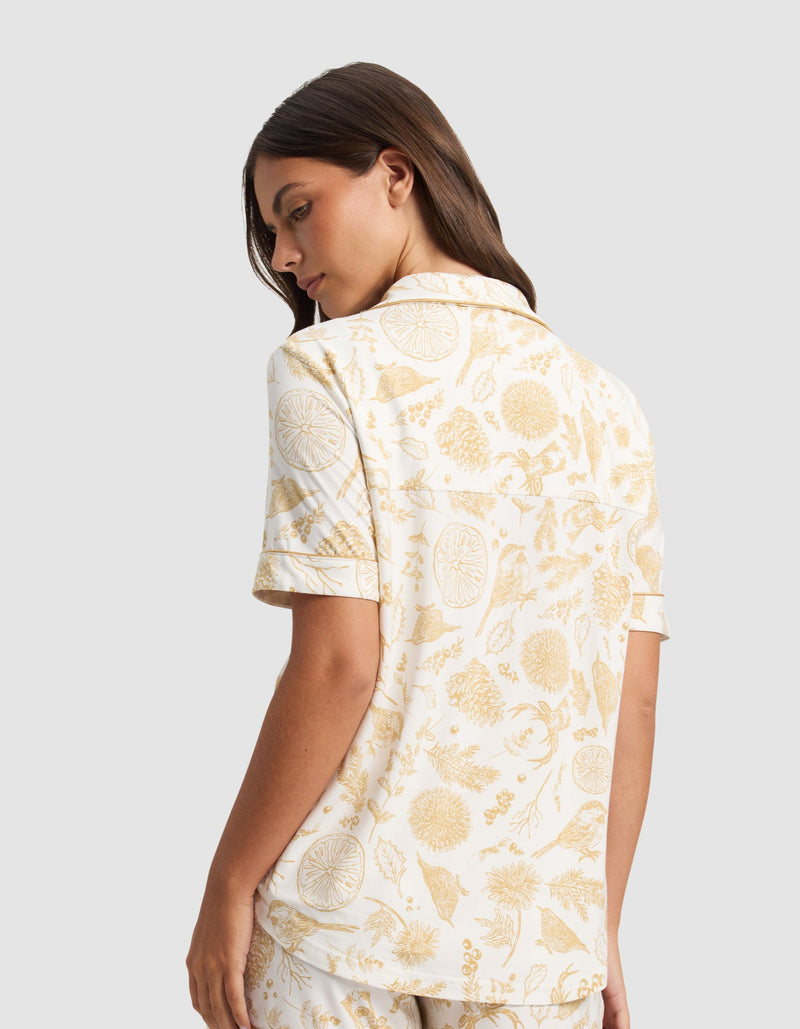 A woman with long brown hair stands turned to the side, wearing the Cozy Earth Women's Bamboo Stretch-Knit Short Sleeve Pajama Top featuring a yellow botanical and citrus fruit print on a white background.
