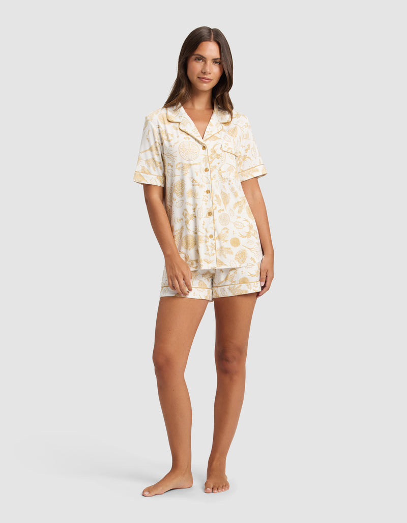 A woman stands barefoot against a light gray background, wearing Cozy Earth's Women's Bamboo Stretch-Knit Short Sleeve Pajama Top and matching shorts with a light yellow and white fruit print.