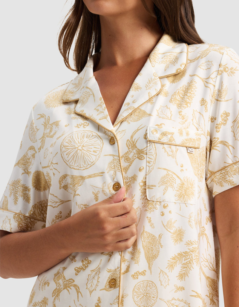 A woman wears the Cozy Earth Women's Bamboo Stretch-Knit Short Sleeve Pajama Top, featuring a white button-up design with gold botanical prints. She holds the shirt's front with one hand. The image is cropped at her shoulders and above the waist.
