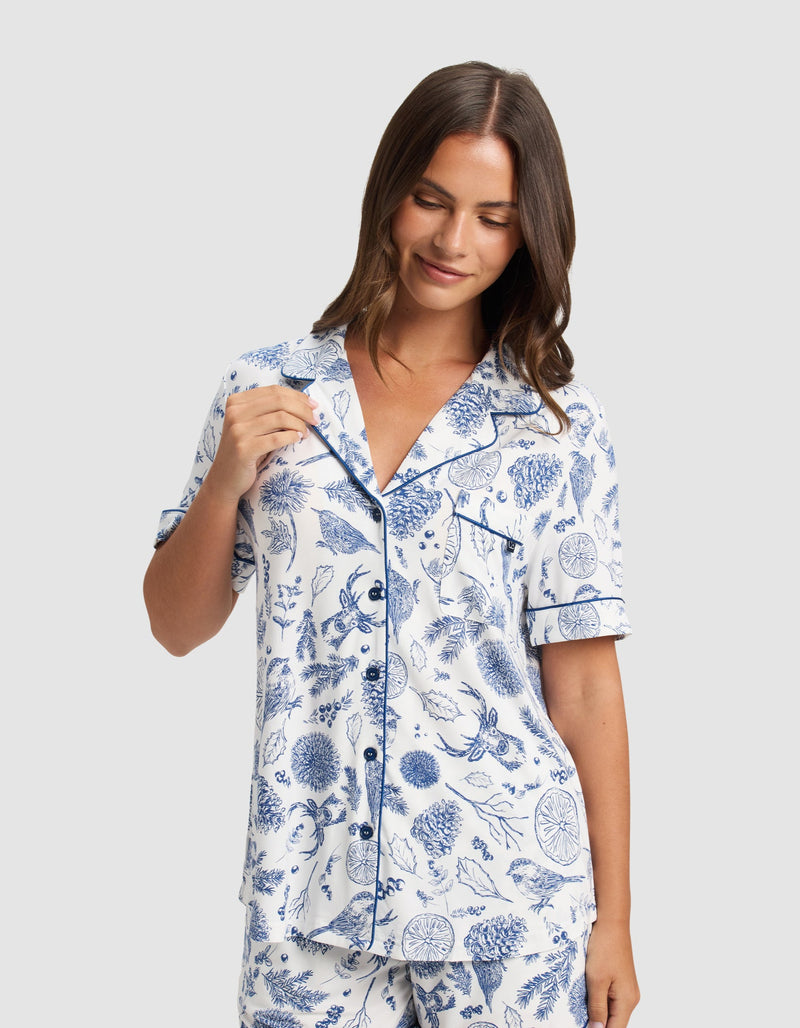 A woman smiles wearing the Cozy Earth Women's Bamboo Stretch-Knit Short Sleeve Pajama Top and matching shorts featuring a blue botanical print on a white background.