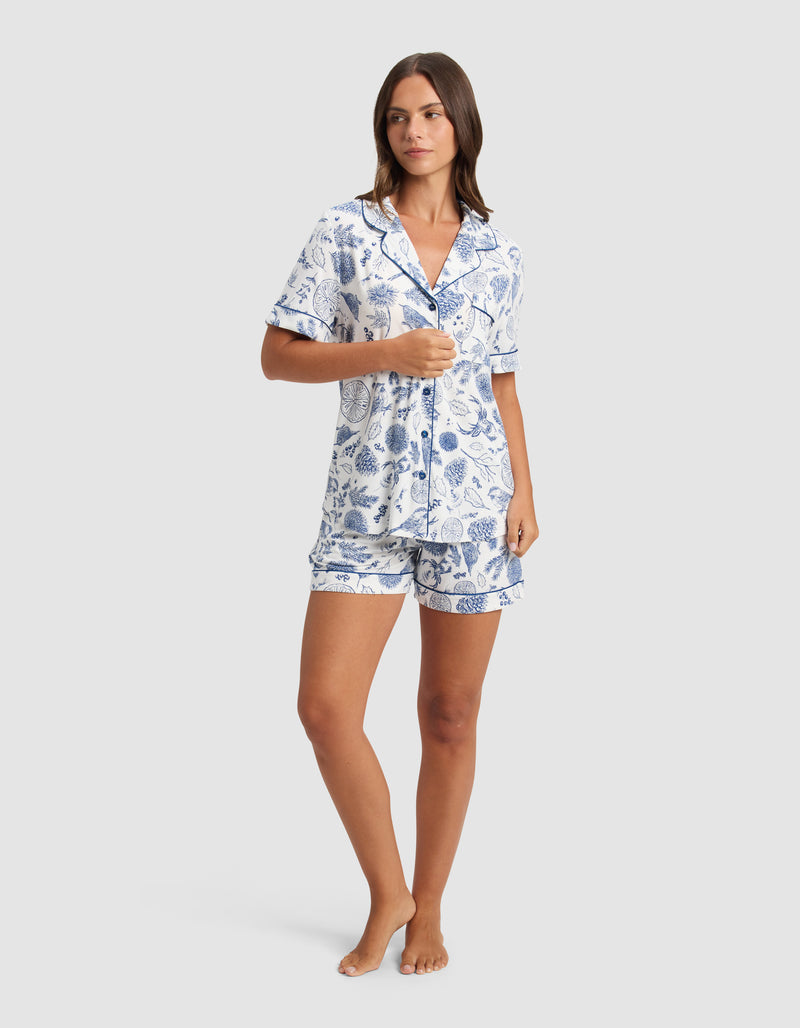A woman with straight brown hair and a neutral expression stands barefoot wearing the Cozy Earth Women’s Bamboo Stretch-Knit Short Sleeve Pajama Top, featuring buttons, a collar, and a blue and white floral pattern with coordinating mid-thigh shorts.