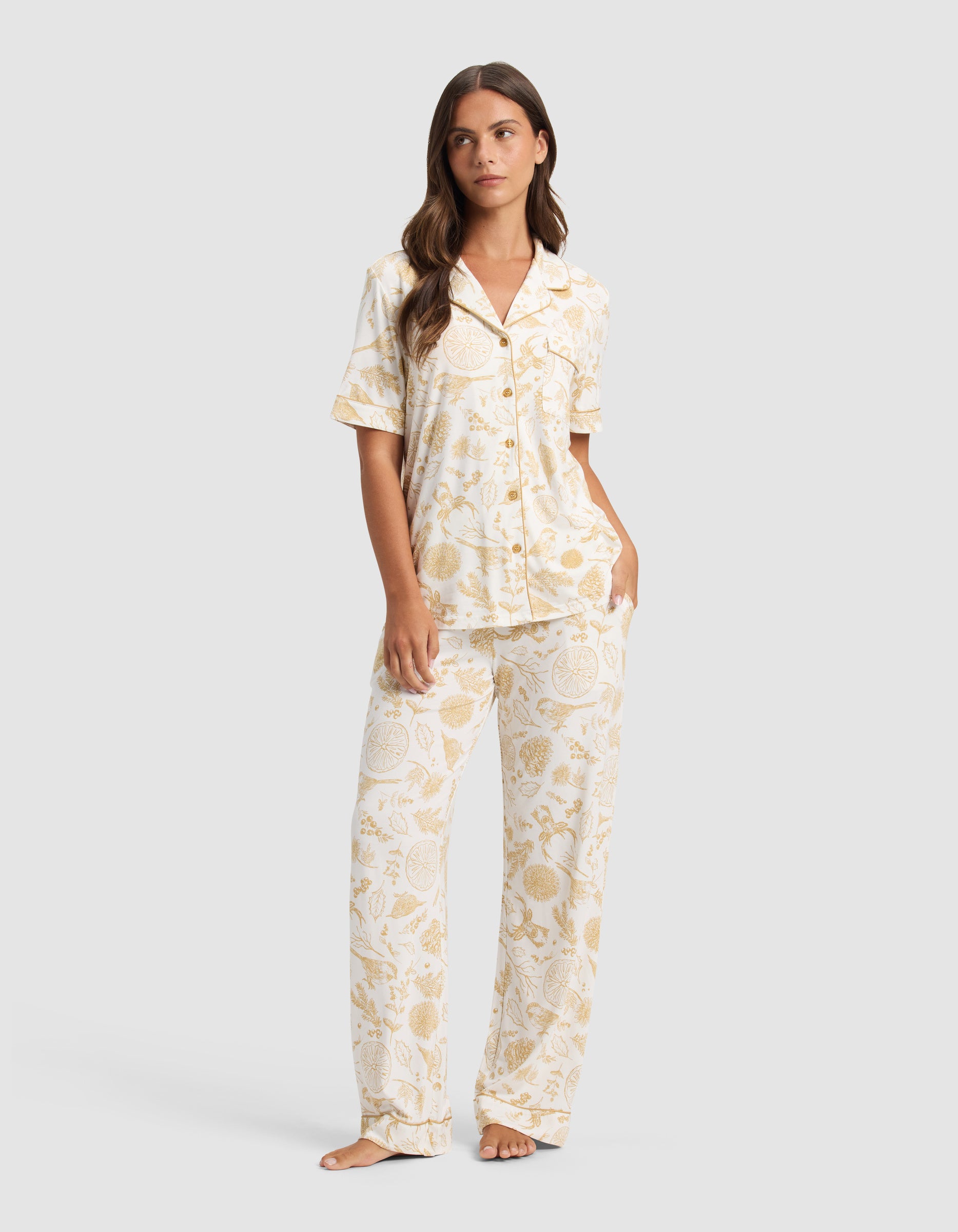 A woman with long brown hair stands barefoot, wearing Cozy Earth's Women's Bamboo Stretch-Knit Short Sleeve Pajama Set in a light yellow and white botanical print. She looks slightly to the side.