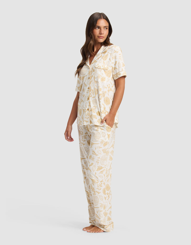 A woman stands barefoot in the Cozy Earth Women's Bamboo Stretch-Knit Short Sleeve Pajama Set, featuring a cream and light yellow floral print with short sleeves and long pants, gazing slightly to the side against a plain background.
