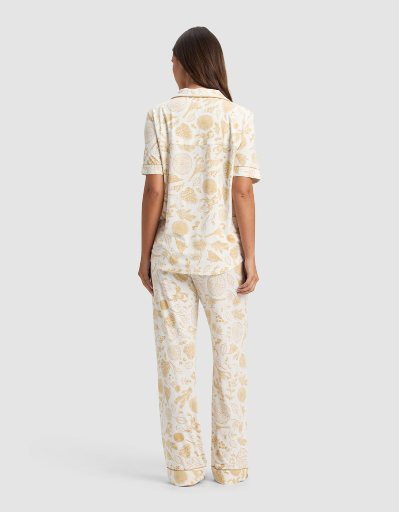 A woman stands facing away, wearing the Cozy Earth Women's Bamboo Stretch-Knit Short Sleeve Pajama Set featuring a light beige botanical print on a white background.